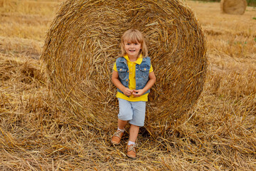Naklejka na ściany i meble a little blond boy with long hair stands by a stack of himself in a mown field