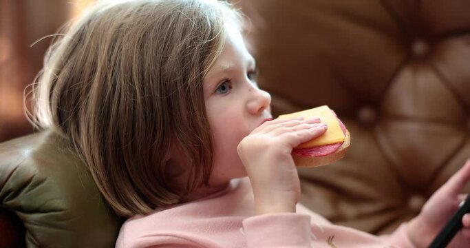 Child lying on sofa and eating delicious sandwich with sausage and cheese 4k movie slow motion. Proper nutrition in children concept