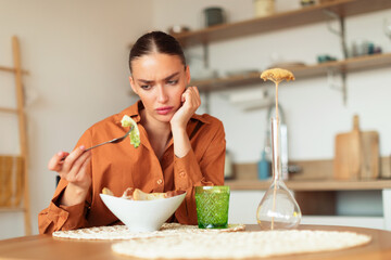 Sad young caucasian lady looking at her caesar salad and dont want to eat, sitting at kitchen at...