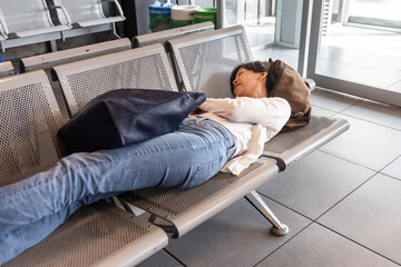 Tired Asian woman lie down sleeping flat on airport departure waiting area chairs while waiting for...