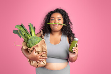 Funny black plus size lady in sportswear with vegetable bag and bottle of smoothie, having fun on pink studio background