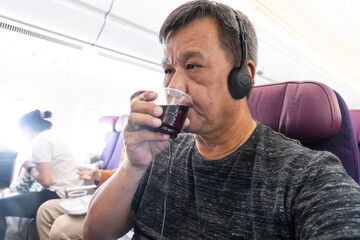 Asian man drinking red wine whilst enjoying inflight entertainment during airplane journey