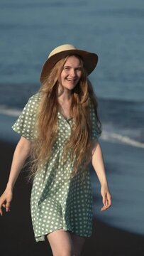 Happy smiling young adult woman model in summer polka dot dress and straw-hat walking on beach of Pacific Coast. Caucasian blonde model looking at camera. Handheld shot. Vertical slow motion video
