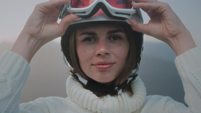 A beautiful Caucasian girl puts a white ski or snowboard helmet on her head. Portrait of a pretty woman at a winter resort. The female wears an orange protective mask or red glasses over her eyes.