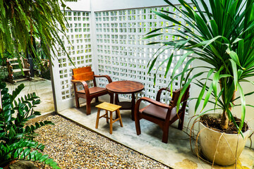 Vintage round wood table and two chair at corner in ornamental garden beside white concrete hole block wall background and tree in flowerpot decoration.