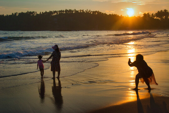 A woman makes a photo of mother and little daughter on the beach at sunset.