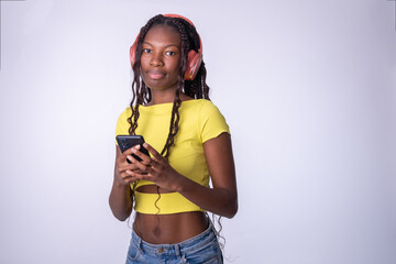 Mujer hermosa joven de color que usa el teléfono inteligente en una fondo blanco. Chica estudiante sonriente enviando mensajes de texto en el teléfono móvil con los auriculares puestos. - obrazy, fototapety, plakaty