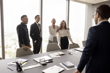 Cheerful business team and business leader standing at large meeting table in boardroom, talking, laughing, enjoying teamwork. Elder businesswoman introducing new younger employee to colleagues