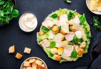Classic Caesar salad with chicken, iceberg salad, croutons, parmesan cheese and caesar dressing. Black table background, top view