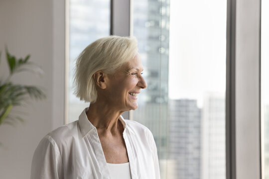Happy blonde senior woman looking at city view out of window, standing indoors, smiling, laughing, thinking, enjoying retirement, leisure, feeling good in comfortable home apartment