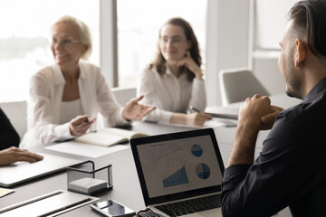 Cropped shot of young business professional man speaking to happy colleagues, partners, telling marketing report on laptop, sitting at meeting table. Businessman with coworkers on blurred background