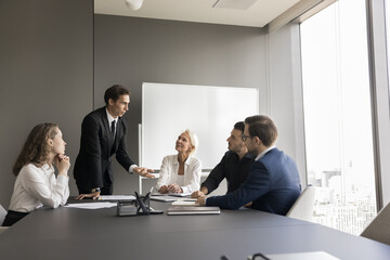 Handsome young business leader man standing at meeting table in boardroom, talking to sitting listening employees. Corporate teacher training work group. Team discussing tasks, planning strategy