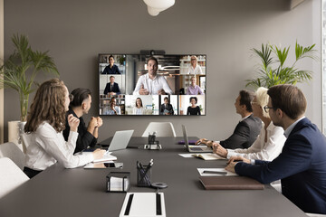 Business teem of office employees and freelancers meeting on online video chat, conference call. Coworkers sitting at negotiation table, looking at display with speaking distance colleagues - obrazy, fototapety, plakaty