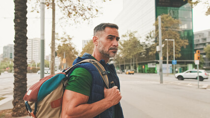 Mature man tourist wearing casual clothes, with backpack on his shoulder, looks around while standing on crosswalk