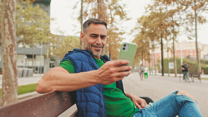 Close-up,happy mature man wearing casual clothes sitting on bench looking through information on smartphone