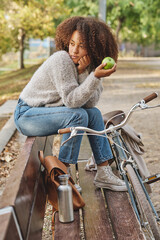 Pensive ethnic woman sitting on bench in park with apple