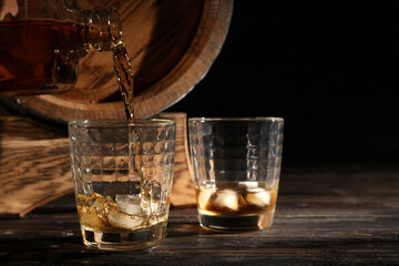 Pouring of cold whiskey from bottle into glasses on dark wooden background