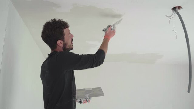 Caucasian working man plastering ceiling to improve and remodel white surface on indoor drywalls in house construction site, handyman with palette knife renovation project, domestic interior