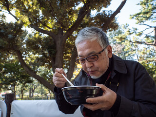 An older asian man eating noodles and soup with chopsticks - 629243101