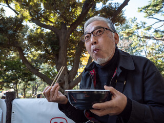An older asian man eating noodles and soup with chopsticks - 629243183
