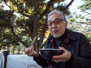 An older asian man eating noodles and soup with chopsticks - 629243301