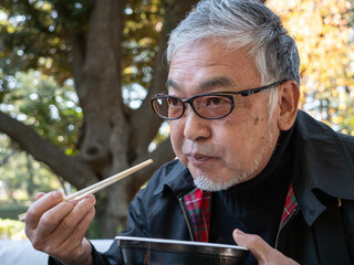 An older asian man eating noodles and soup with chopsticks - 629244114