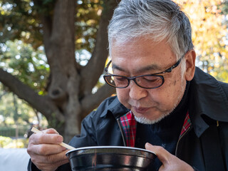 An older asian man eating noodles and soup with chopsticks - 629244393