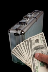 Person's hand grasping a pile of cash dollars beside a silver case, symbolizing financial success © Vardges Petrosyan/Wirestock Creators