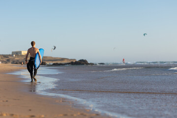 Fit man walking along the shore holding a surfboard - view from the back - kitesurfers on the...