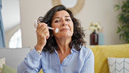 Middle age hispanic woman massaging face with skin roller sitting on sofa at home