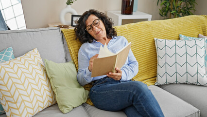 Middle age hispanic woman reading book sitting on sofa at home
