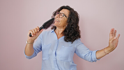 Middle age hispanic woman singing song using brush as a microphone over isolated pink background