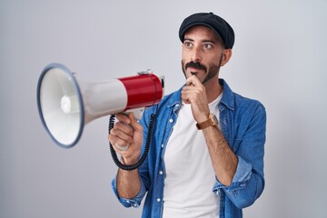 Hispanic man with beard shouting through megaphone serious face thinking about question with hand...