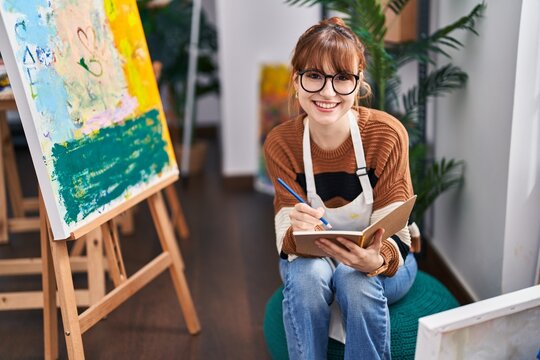 Young woman artist smiling confident writing on book at art studio