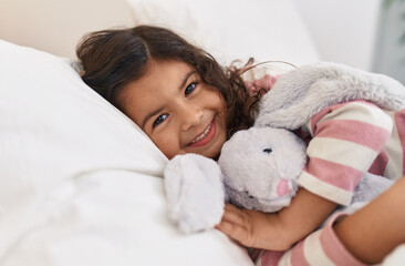 Adorable hispanic girl hugging doll lying on bed at bedroom