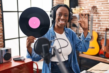 Beautiful black woman holding vinyl record at music studio doing ok sign with fingers, smiling friendly gesturing excellent symbol