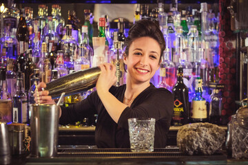 Cheerful female bartender mixing cocktail in shaker at counter in nightclub
