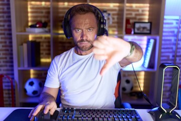 Middle age man with beard playing video games wearing headphones looking unhappy and angry showing rejection and negative with thumbs down gesture. bad expression.