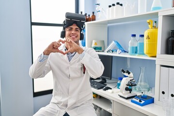 Young hispanic man working at scientist laboratory wearing vr glasses smiling in love doing heart...