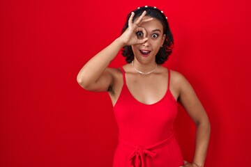 Young hispanic woman standing over red background doing ok gesture shocked with surprised face, eye looking through fingers. unbelieving expression.