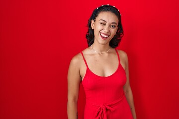 Young hispanic woman standing over red background winking looking at the camera with sexy expression, cheerful and happy face.