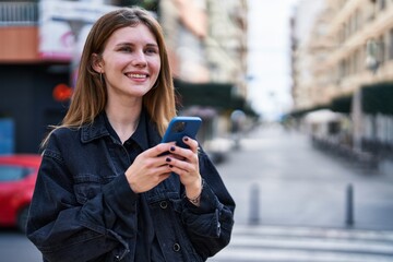 Young blonde woman using smartphone smiling at street