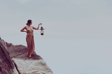 Mature woman stands on the side of a mountain, with an antique lantern holding in her hands © Underapalmtree/Wirestock Creators