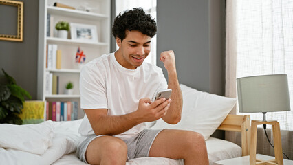 Young latin man using smartphone sitting on bed celebrating at bedroom