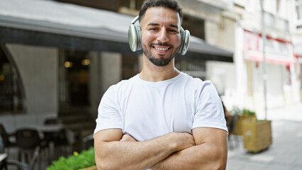 Young arab man listening to music standing with arms crossed gesture at coffee shop terrace