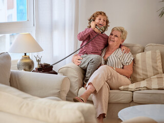 Laughing boy answering telephone call while resting with aged grandmother