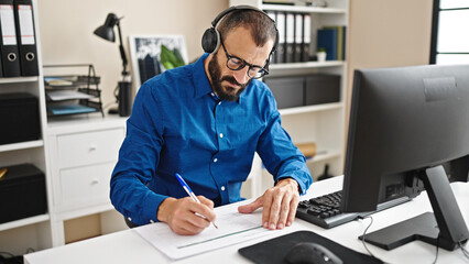 Young hispanic man business worker using computer and headphones taking notes at office