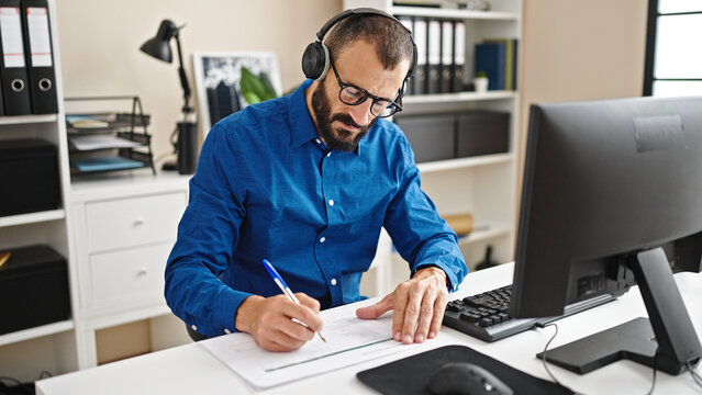 Young hispanic man business worker using computer and headphones taking notes at office