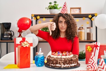 Hispanic woman with curly hair celebrating birthday holding big chocolate cake with angry face, negative sign showing dislike with thumbs down, rejection concept