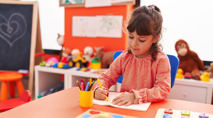 Adorable hispanic girl student sitting on table drawing on notebook at kindergarten